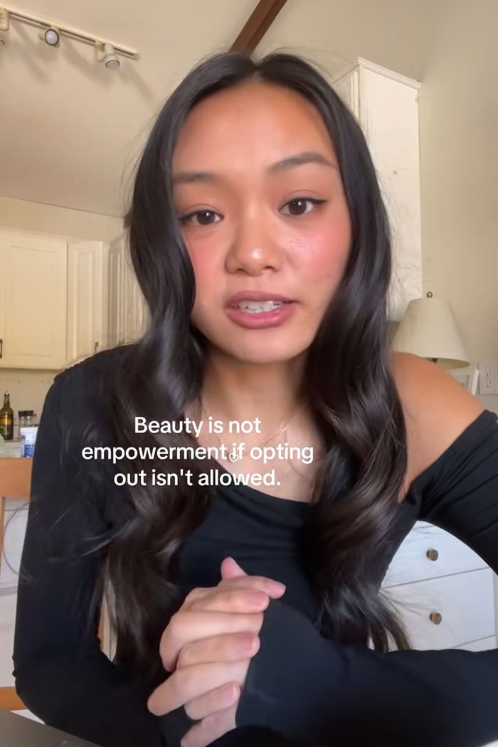 Young woman explaining beauty standards and empowerment in a casual indoor setting with long dark hair and a black top.