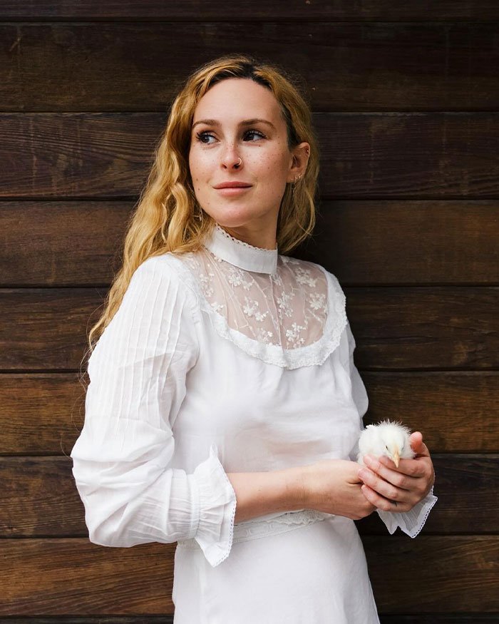 Rumer Willis wearing white holding a small chick, standing against a wooden wall, highlighting work and famous parents.