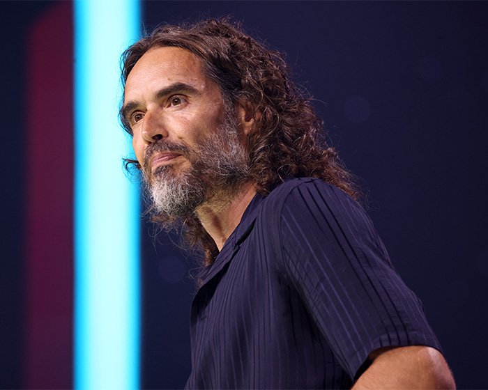Russell Brand with long hair and beard, speaking at an event amid controversy and disturbing charges news.