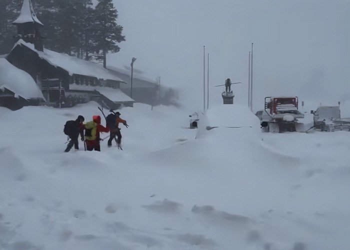 Six mothers in winter clothing walking through deep snow near a snowy village scene after enjoying time off together.