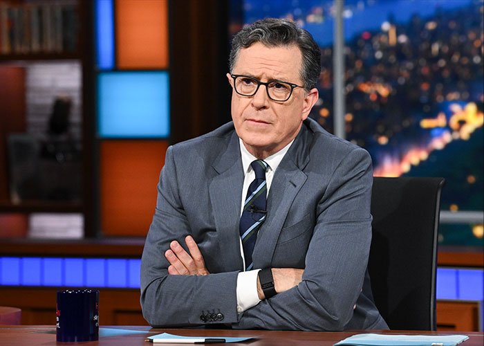 Stephen Colbert wearing a suit and glasses with arms crossed on a late night show set, responding to Epstein Files news.