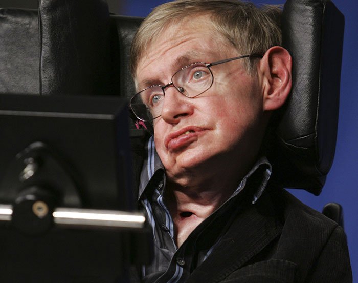Stephen Hawking sitting in a motorized chair wearing glasses and a dark jacket during a public appearance.