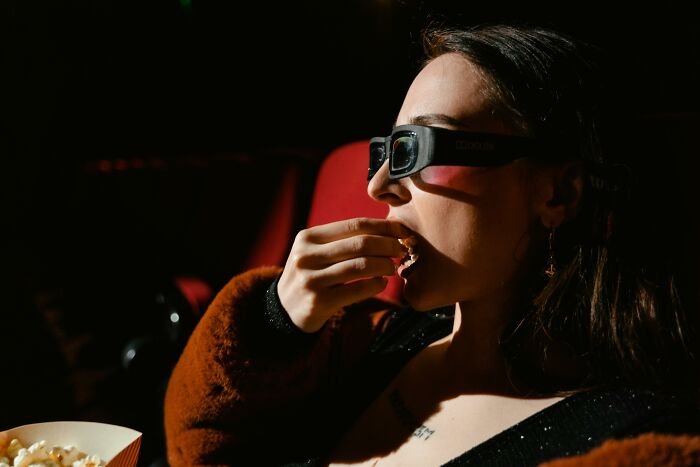 Woman wearing 3D glasses eating popcorn in a dark theater, enjoying a movie related to Tom Hanks side character quiz.