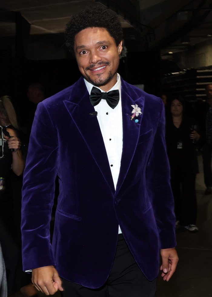 Trevor Noah smiling in a purple velvet jacket and bow tie at a Grammys event backstage with people in the background