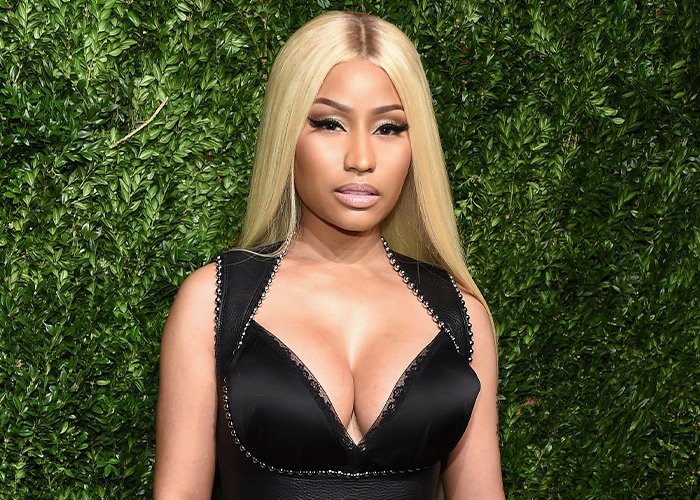 Nicki Minaj with dramatic new look, long blonde hair and black outfit, posing against green leafy background.