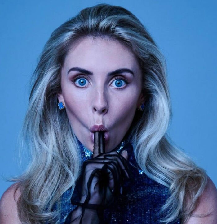 Blonde woman with blue eyes wearing black gloves and blue earrings, making a shushing gesture in a blue-toned setting.