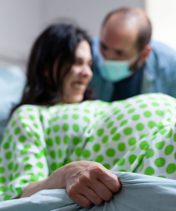 Woman in labor wearing green polka dot gown gripping bedsheet, man with mask supporting her during childbirth moment.
