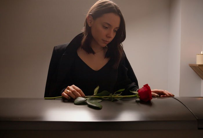 Woman in black mourning outfit holding a red rose, reflecting on inheritance from her late husband in a dim room.