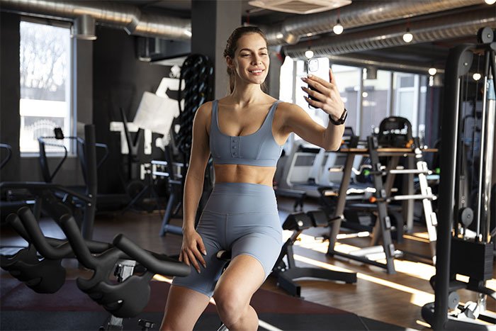 Woman at the gym taking a selfie with a phone, wearing workout clothes in a modern fitness center.
