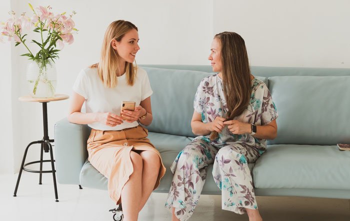 Two women sitting on a couch, one holding a phone, appearing surprised by roommates parents using a spare key.