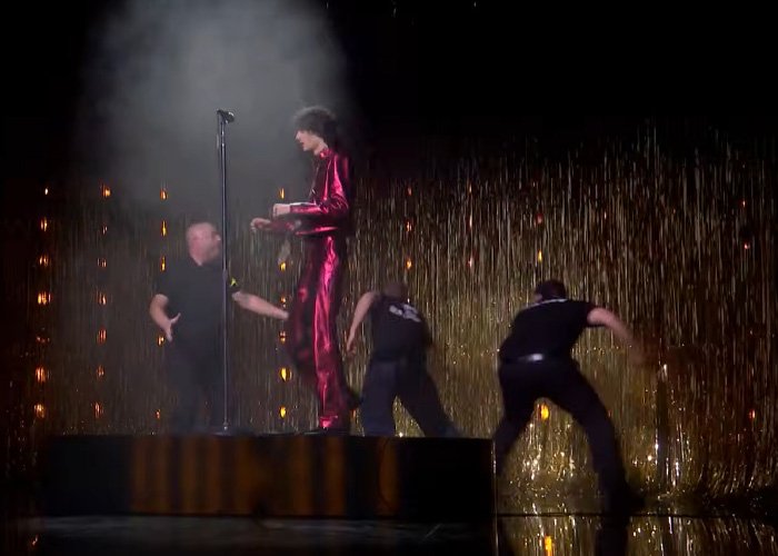 Singer in red outfit being pushed off stage during chaotic Brit Awards performance with security and gold curtain backdrop.