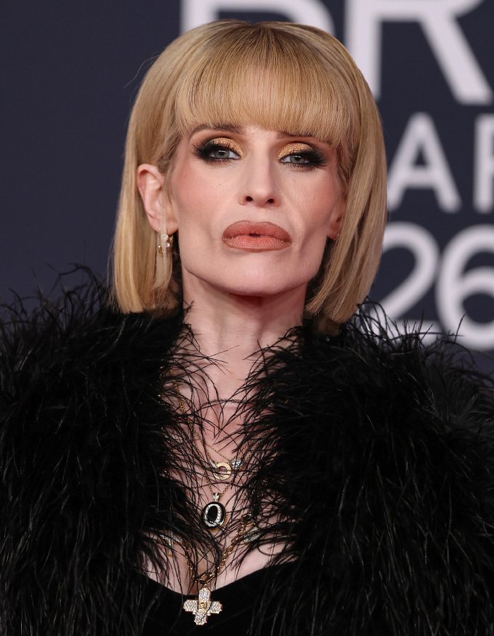 Kelly Osbourne wearing a black feathered outfit and layered necklaces at the Brit Awards with bold makeup and a blunt bob hairstyle.