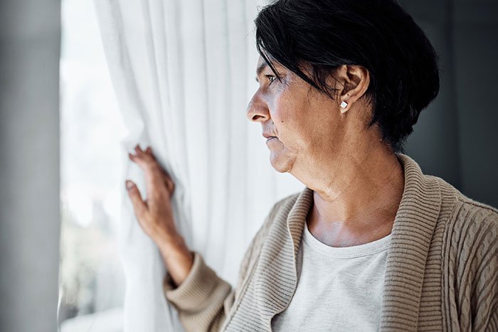 Man stressed and paranoid looking out window, worried about unhinged neighbor not leaving him alone at home.